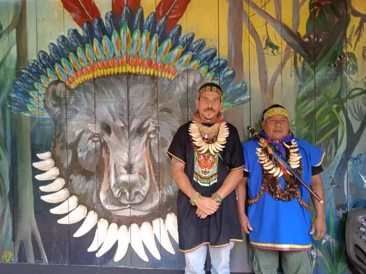 Gabriel Guillén and Taita Alirio Causaluzán in ceremonial regalia in front of the bear-spirit mural