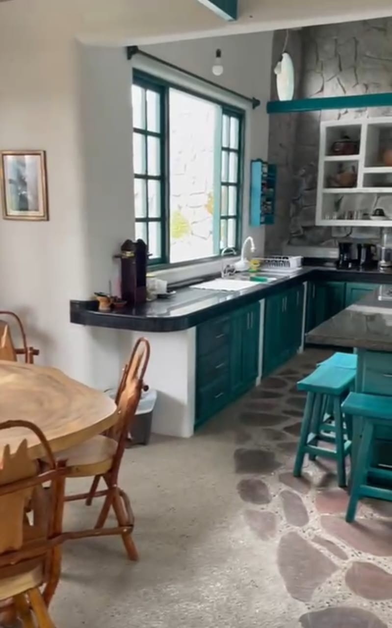 The communal kitchen at Teos — teal cabinets, stone wall, dining nook with handcrafted wooden chairs